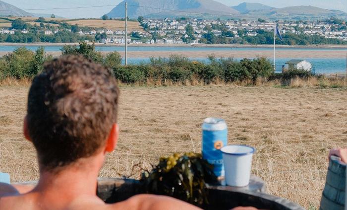 A man bathing at the Wild Môn Seaweed Baths at Halen Môn, in a warm 40C barrel with a view of Eryrion a sunny day with a beer